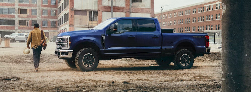 A side view of a blue 2026 Ford F-250 pickup parked at a work site, with a person in a brown jacket walking away from the truck