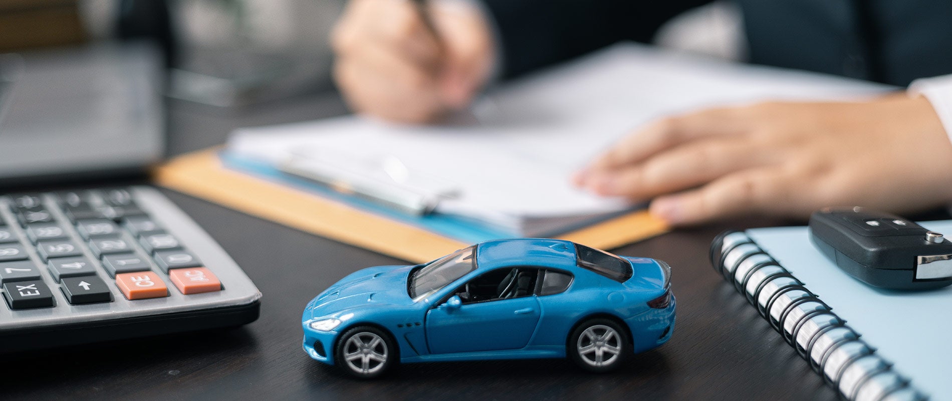 The hands and arms of a white person in a suit, sitting at a desk with a notepad, calculator, key fob, and blue toy car