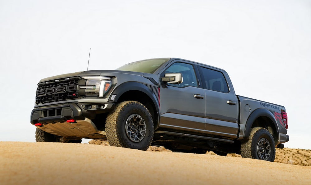The hands and arms of a white person in a suit, sitting at a desk with a notepad, calculator, key fob, and blue toy car